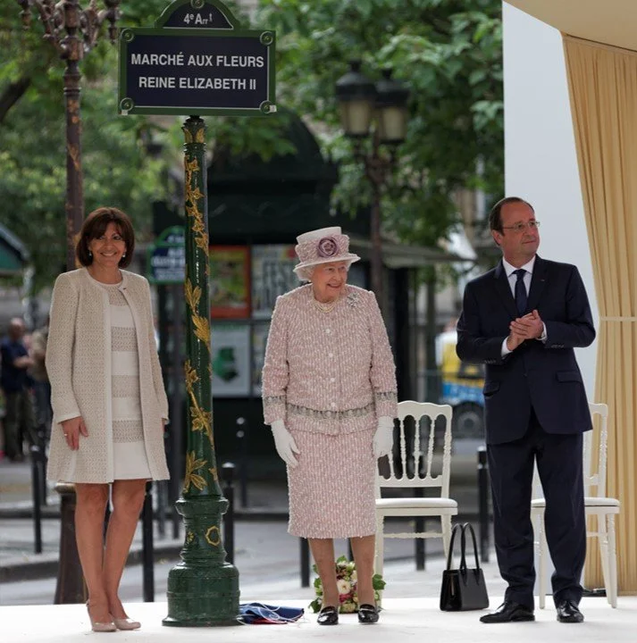 Queen Elizabeth II in a pink outfit and hat standing on a stage during the Paris Flower Market, with two other people, one woman and one man, standing beside her.