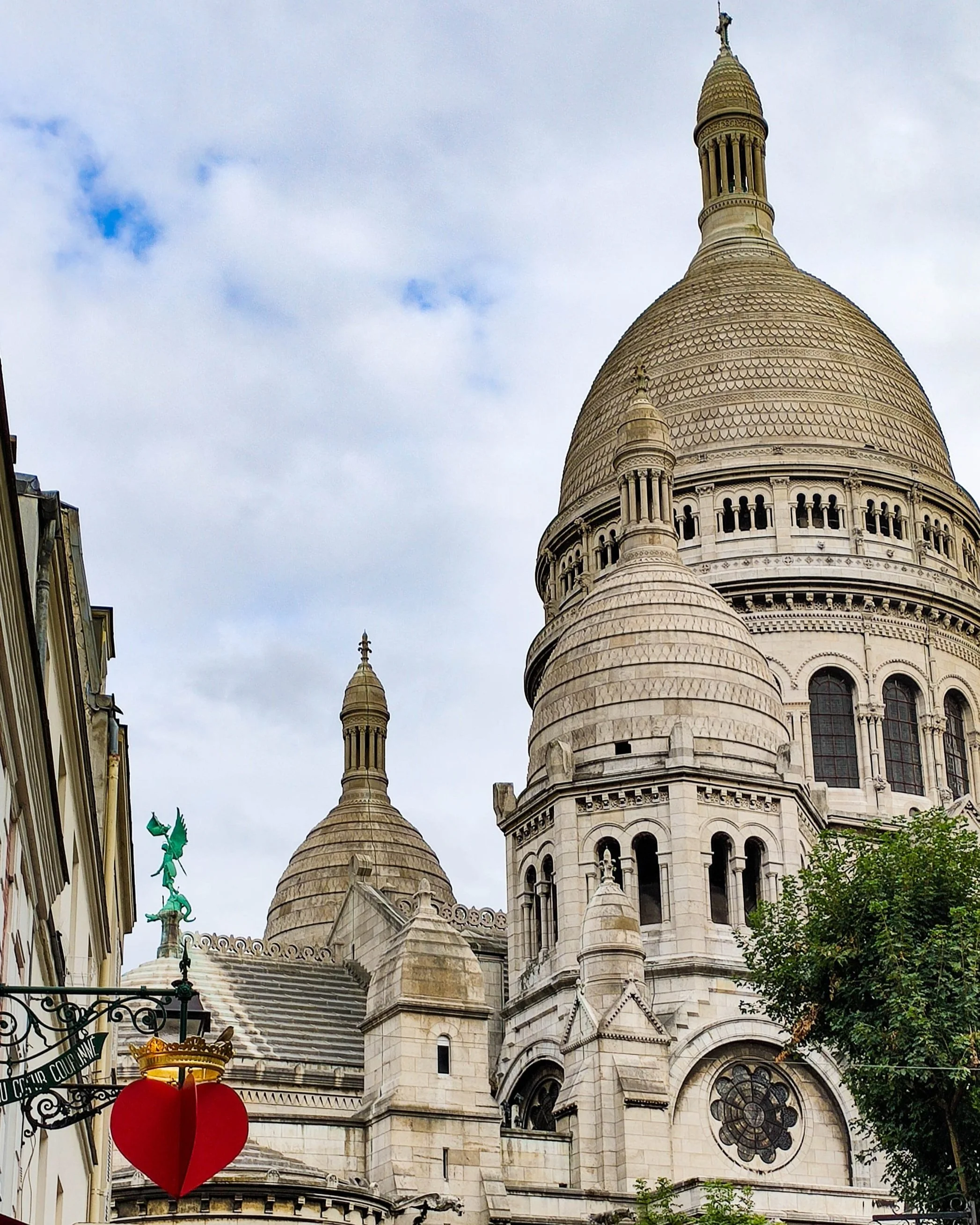 View of the Sacré-Cœur Basilica in Paris, France, showing its domes, detailed architectural features, and a statue of a figure on a rooftop. Part of a city street with trees and buildings in the foreground.
