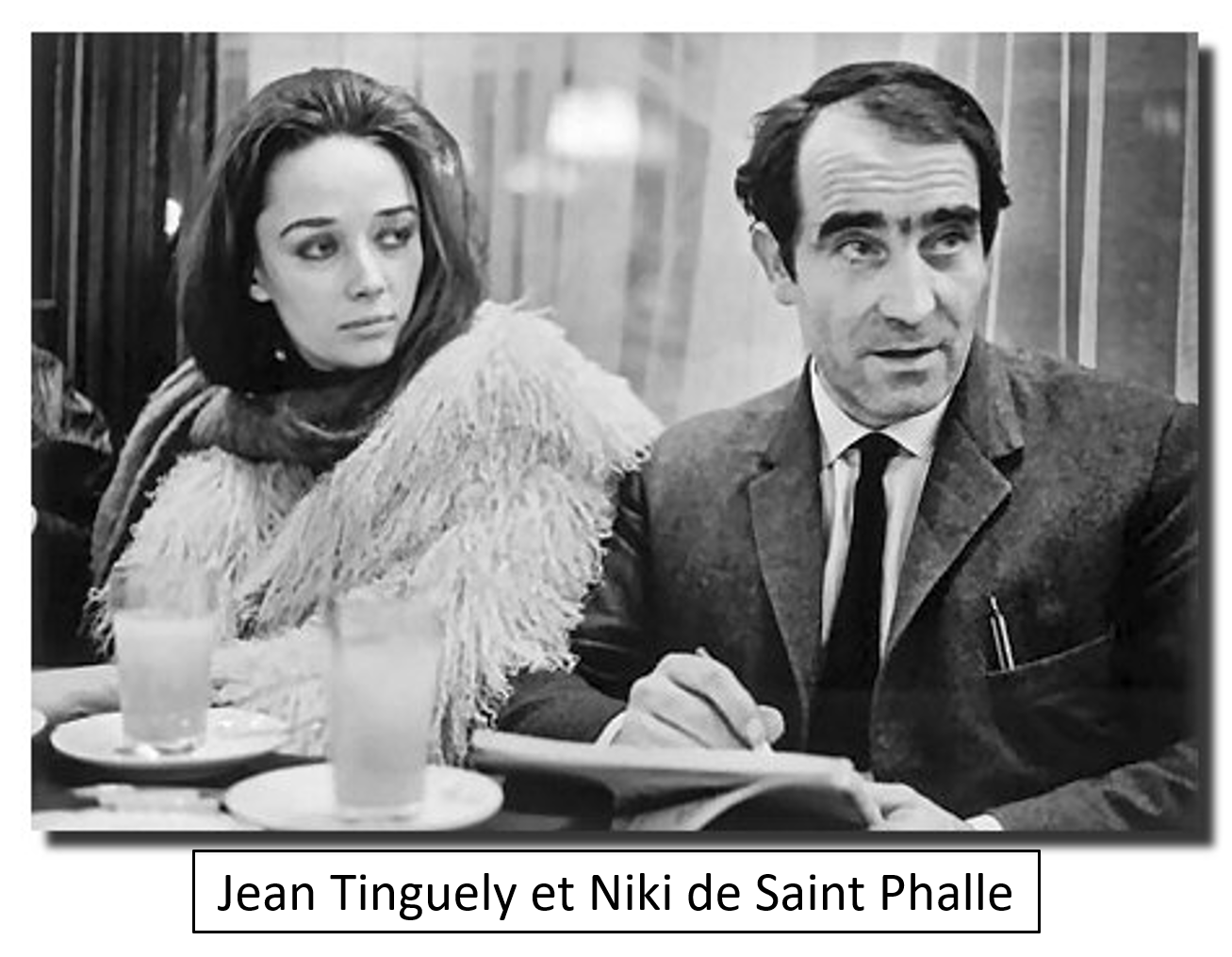 A black-and-white scene from a movie showing a woman with dark hair and a man with dark hair and eyebrows sitting at a table in a restaurant or café. The woman is looking at the man with a serious expression. There are drinks on the table.