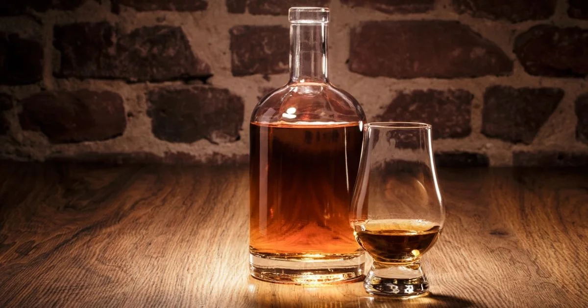 A glass decanter filled with amber liquid on a wooden surface, with a glass of whiskey beside it, against a brick wall background.