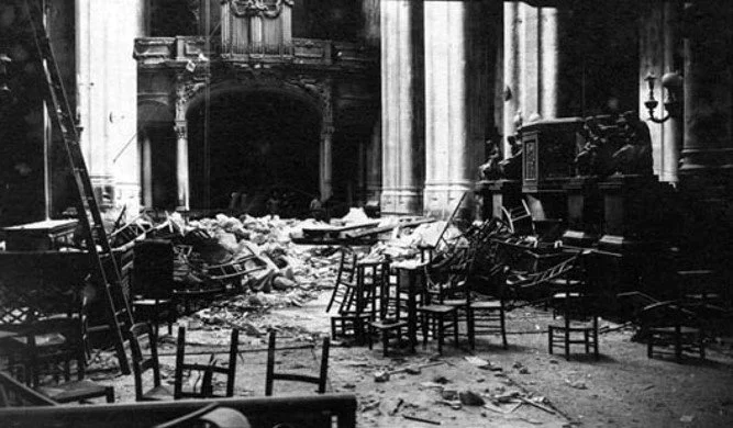 Black and white photograph of a large, historic room with a grand fireplace and high ceilings, showing signs of damage with debris and overturned chairs scattered across the floor.