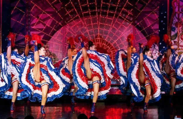 A group of dancers performing a high-kick dance routine on stage, wearing colorful dresses with red, white, and blue ruffles, under a vibrant, illuminated backdrop.
