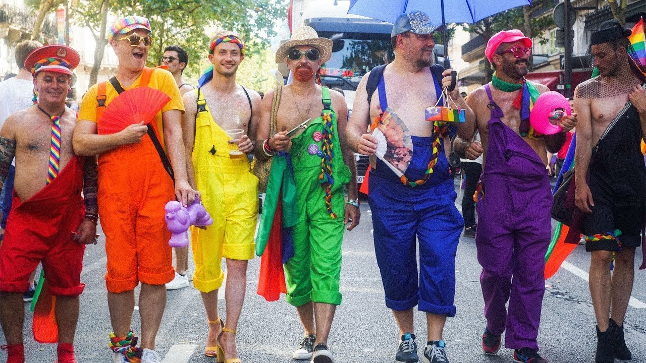 A group of people participating in a pride parade, wearing colorful outfits and accessories that symbolize LGBTQ+ pride. They are walking on a street, some holding rainbow flags, balloons, and drinks, and appear to be celebrating.
