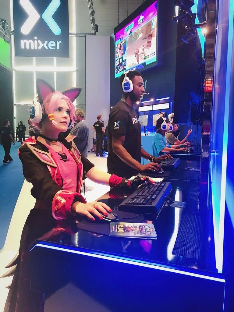 People playing video games at a gaming event, with the 'Mixer' logo visible on the wall, wearing headphones, and using gaming keyboards and mice.