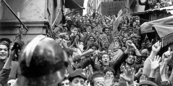 Historical black and white photo of a large crowd of people protesting or demonstrating, with many raising their hands and showing expressions of enthusiasm or determination.