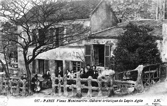 A vintage black and white photo of a group of people outside a building, with a tree in front. The building has open windows with shutters and appears old, with some graffiti on the wall. The scene is from Montmartre, Paris, at an artistic cabaret ca