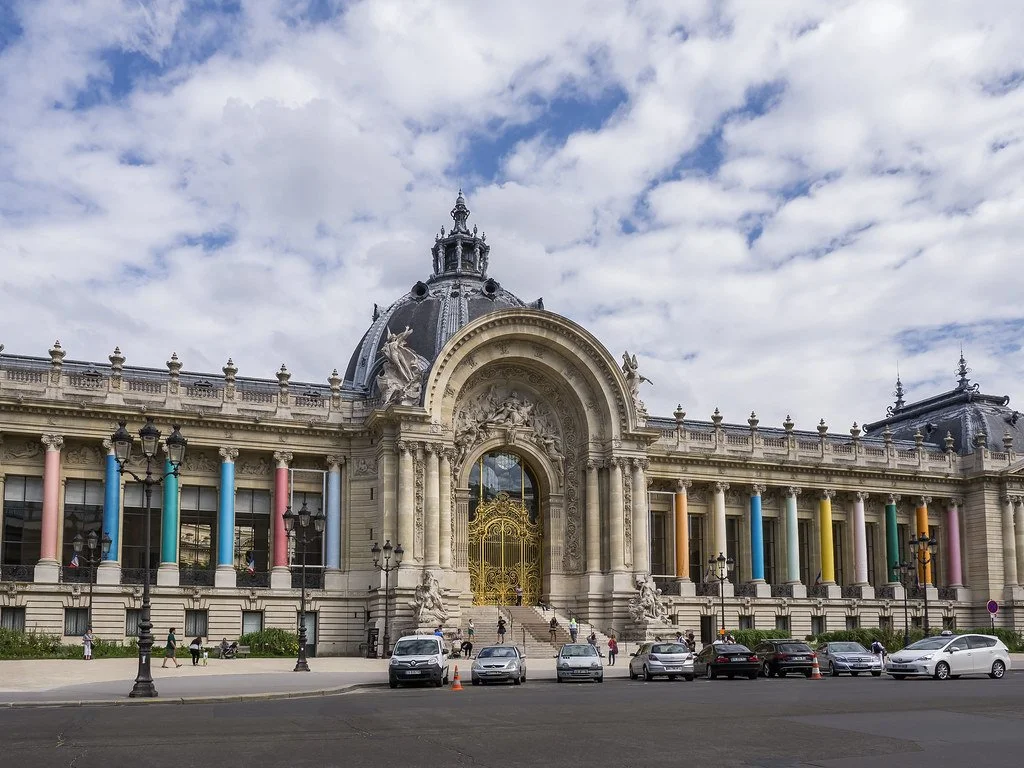 A large historic building with grand architecture, including a domed roof, columns painted in various colors, and ornate sculptures. Cars are parked in front of the building and people are walking nearby.