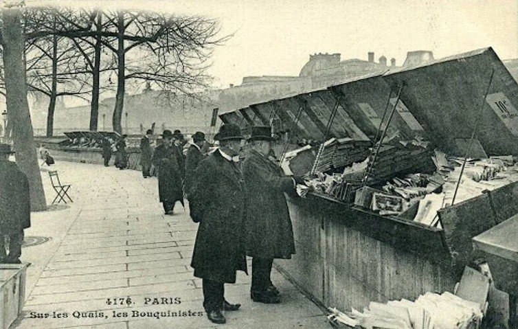 Black and white photo of people standing along a Paris outdoor bookstall at a sidewalk market, with trees and buildings in the background.