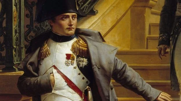 Painting of Napoleon Bonaparte in a military uniform with medals, standing with one arm resting on a surface, inside a grand building with stairs in the background.