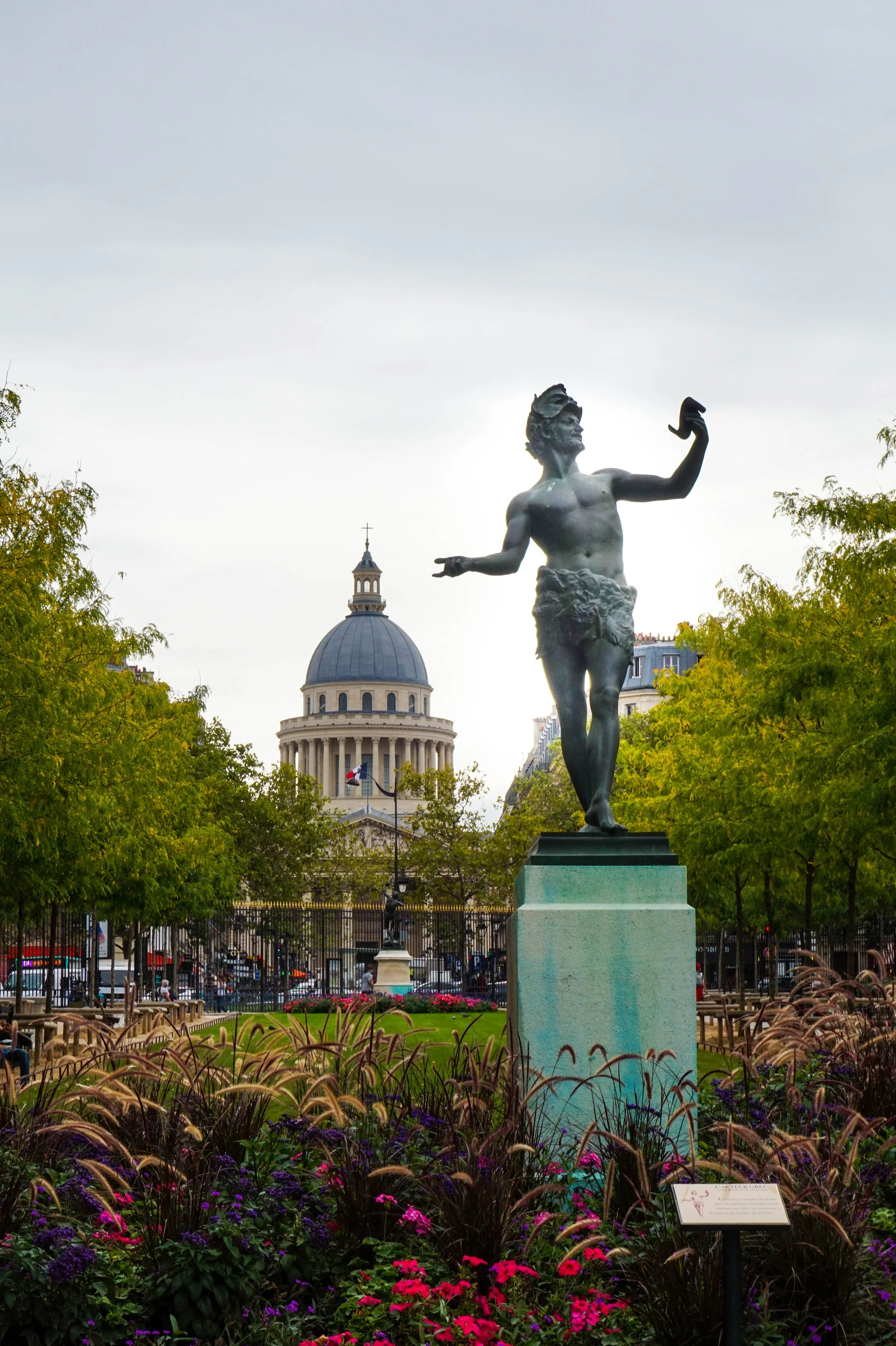 Statue of a man with a beard and crown, shirtless, wearing a skirt, standing on a pedestal in a park with trees and flowers, with a dome building in the background under a cloudy sky.