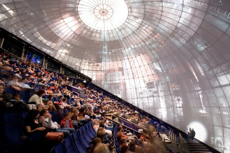An indoor auditorium filled with children seated in blue chairs, watching a performance or presentation on stage, with a large glass dome ceiling overhead.