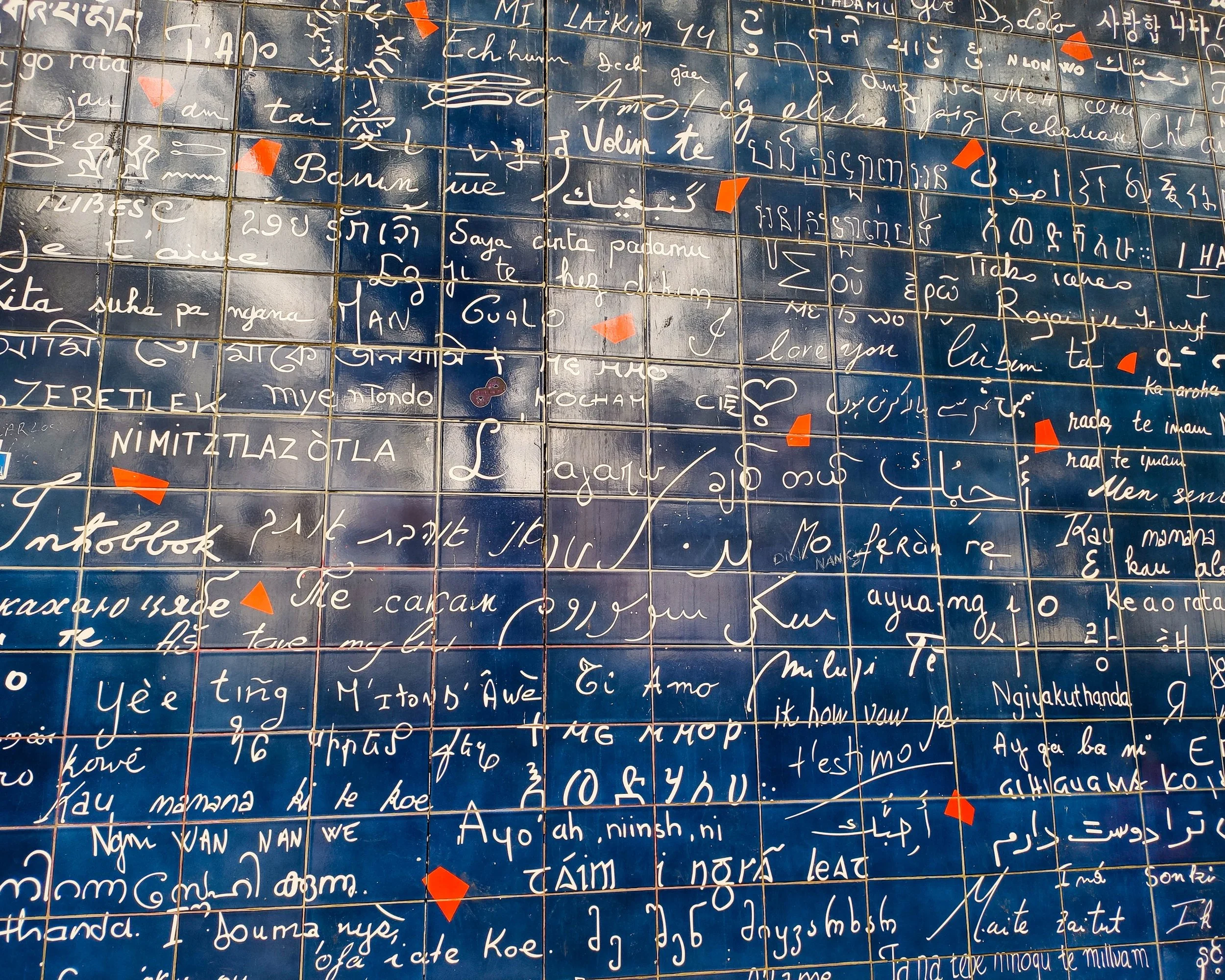 A mosaic wall featuring handwritten messages in various languages on blue tiles, with some tiles having small red arrow stickers.