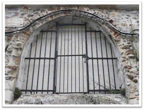 A black metal gate with a padlock, set within an arched stone opening in a brick and stone wall.