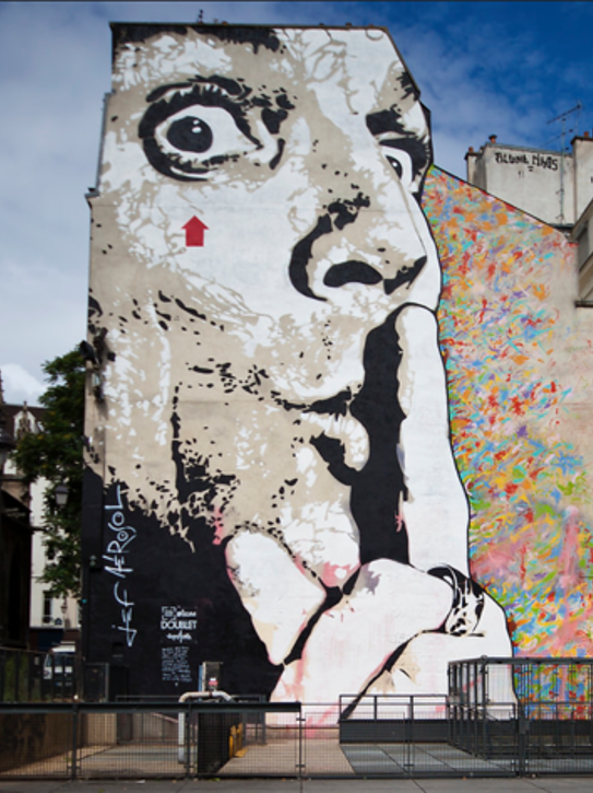 Large mural of a woman's face with finger over lips in a shushing gesture, partially painted in black and white, with colorful abstract background on an urban building wall.