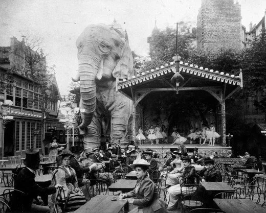 An outdoor cafe scene with many people seated at tables, some wearing vintage clothing and hats, with a large elephant sculpture above a stage where dancers perform. The setting appears to be in a city park or plaza.