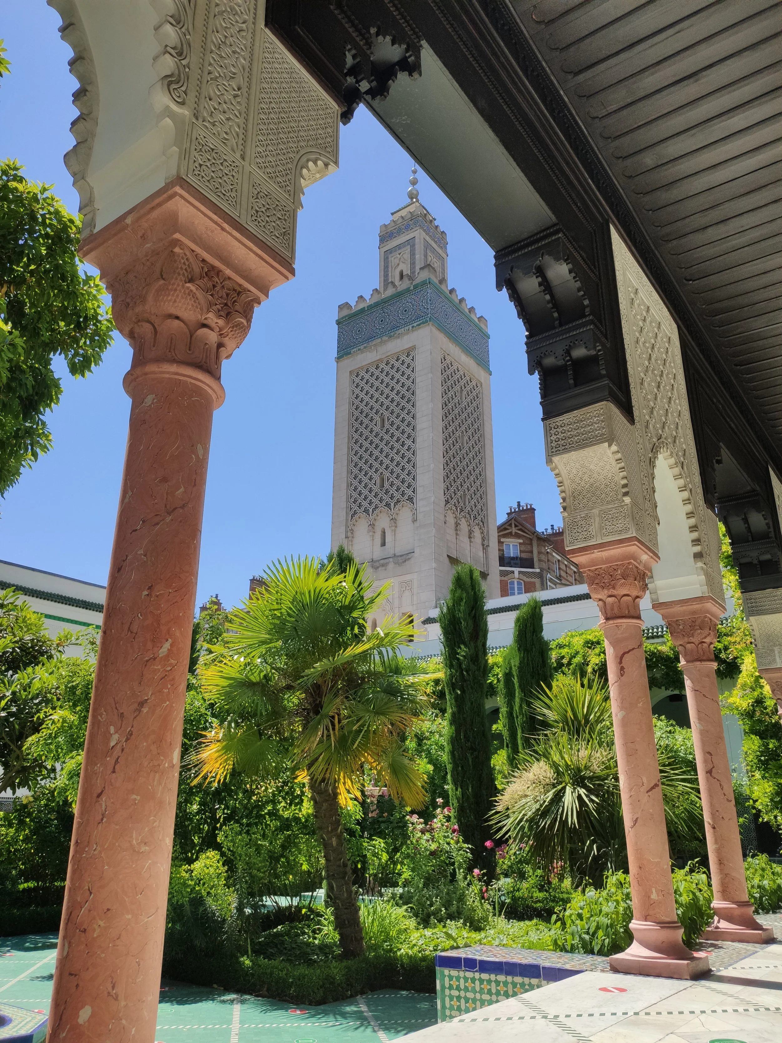 Tourist view of the Hassan II Mosque in Casablanca, Morocco, seen from a courtyard with lush green plants and pink marble columns, under a sunny blue sky.