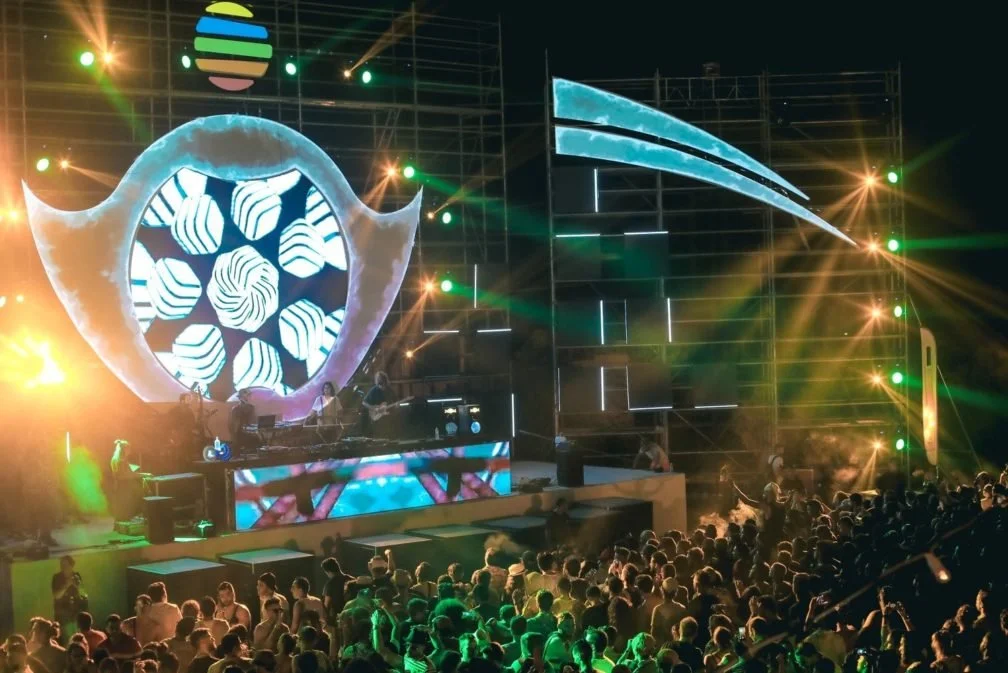 A live outdoor concert with a large crowd, colorful stage lights, a digital backdrop, and a band performing.
