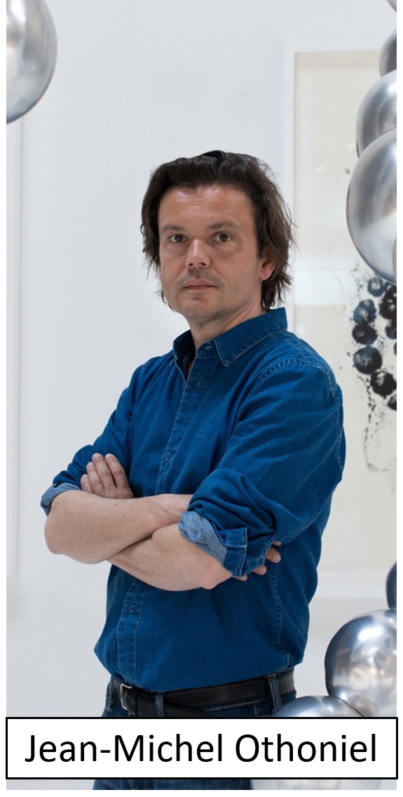 A man with dark, slightly wavy hair wearing a blue denim shirt, standing with arms crossed in front of abstract artwork and metallic balloons.