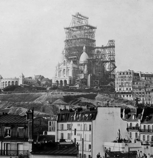 Construction scaffolding around a large cathedral or domed building under renovation, with surrounding city buildings and a hilly landscape in the foreground.