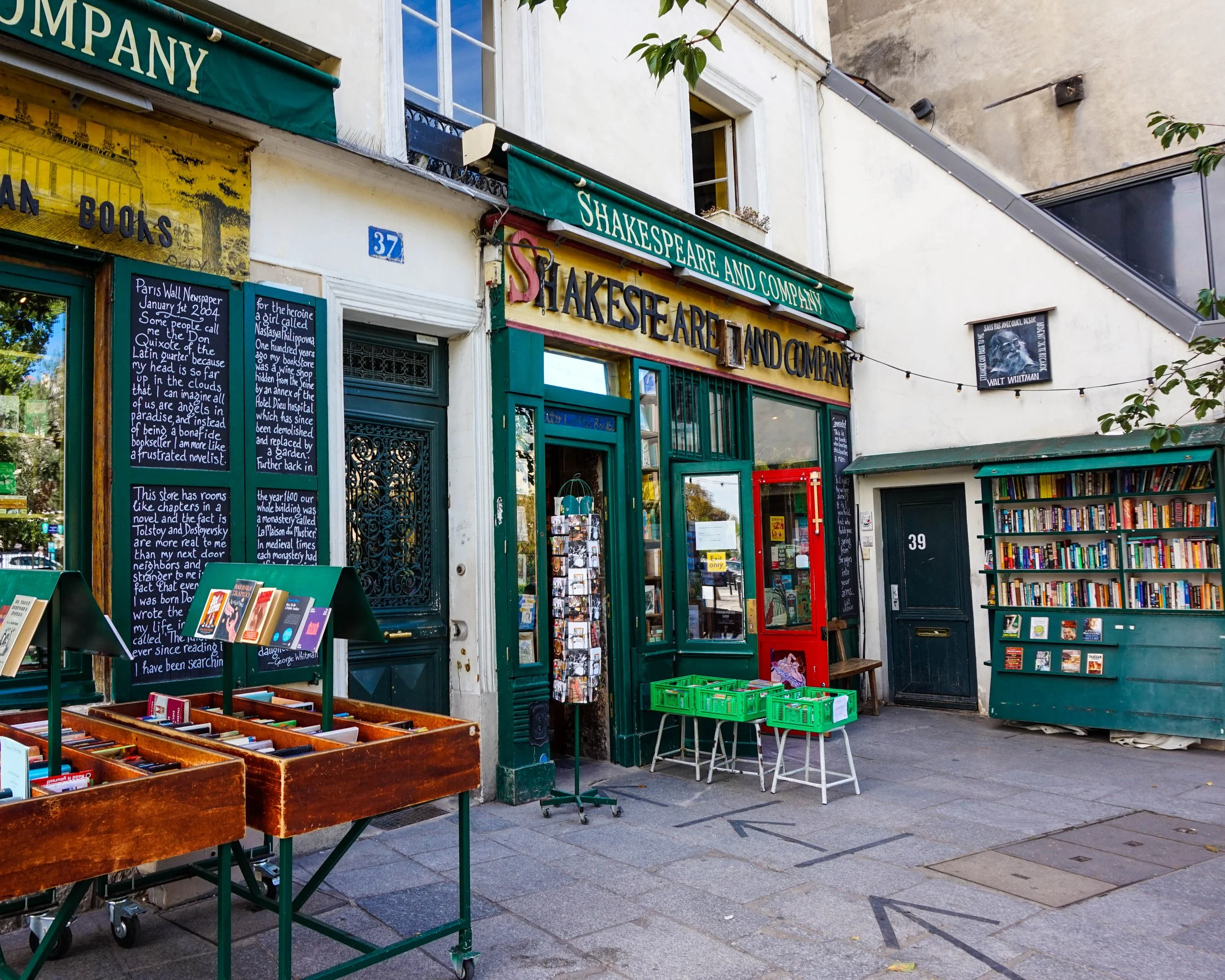 Exterior of a bookstore named 'Shakespeare and Company,' with books displayed outside, a green storefront, blackboards with handwritten notes, and a small outdoor book rack.