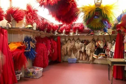 A variety of colorful feather headdresses and costumes hanging on display in a store or costume shop, with shelves and bins visible.