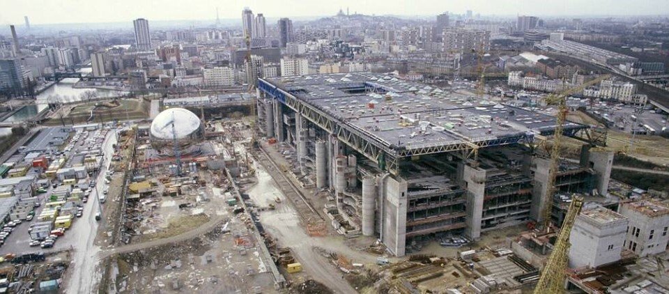 A large construction site with a partially built structure, multiple cranes, and a city skyline in the background.