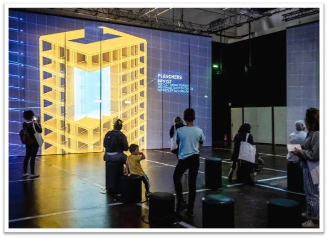People at an indoor exhibition viewing a large digital display of a yellow 3D model of a building structure with blue accents.