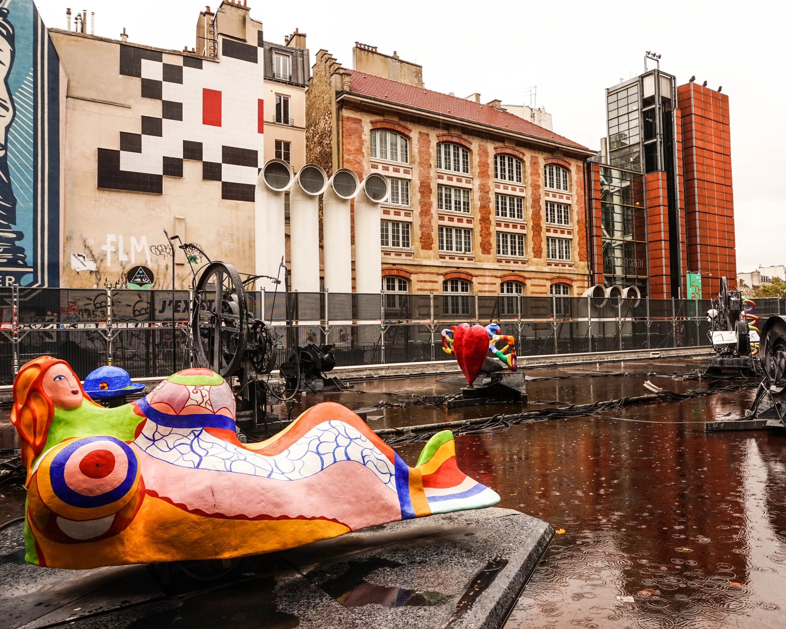 Colorful sculptures of a mermaid and a heart with a rainbow pattern, placed on a wet platform in an urban setting with buildings and street art visible in the background.