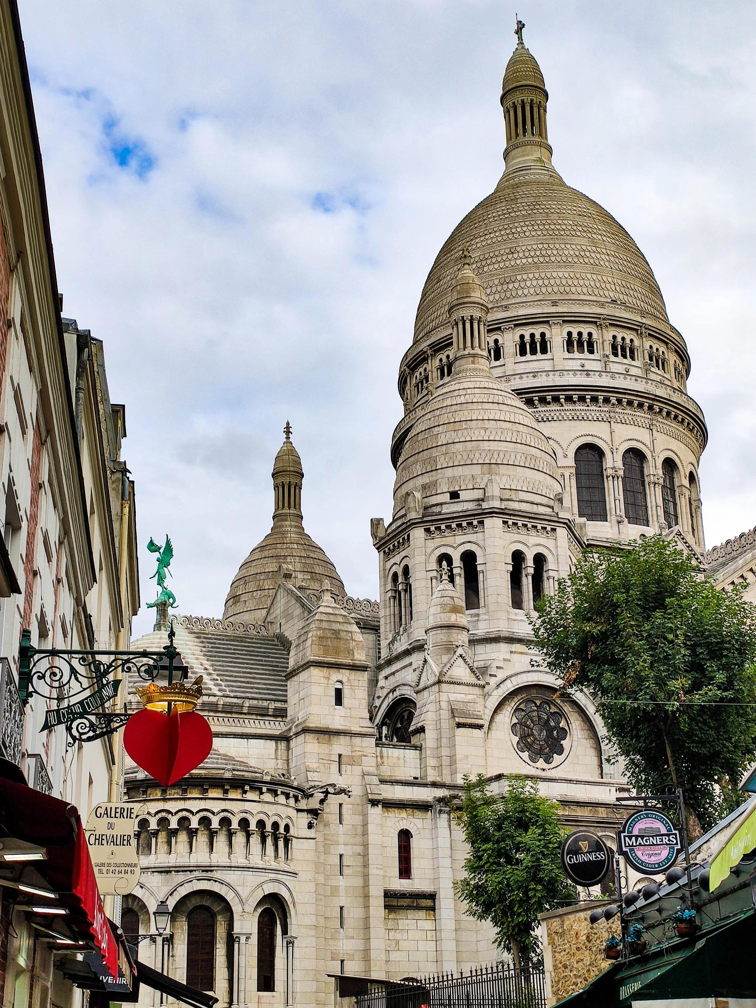 The Sacré-Cœur Basilica in Paris, France, with its large dome, smaller towers, and ornate stonework. Commercial signs and a tree are visible in the foreground.