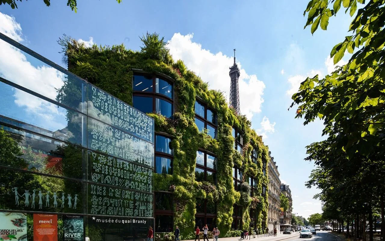 A building covered in green plants and trees on its exterior, with a view of the Eiffel Tower in the background on a sunny day with blue sky.