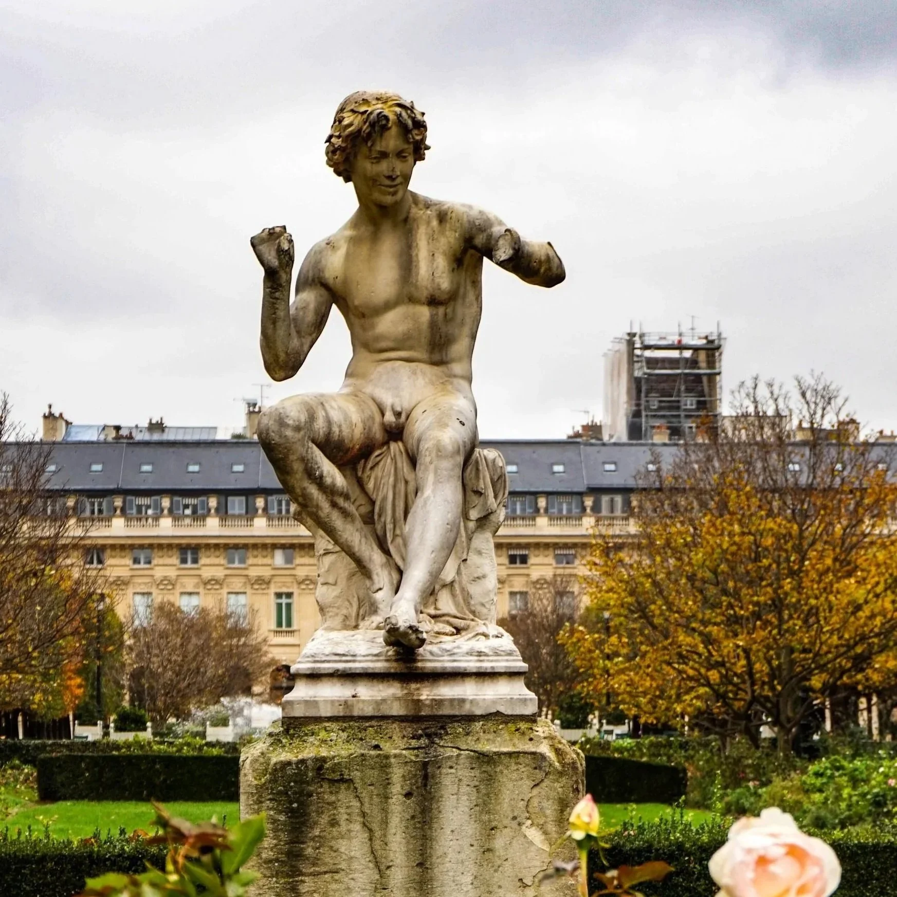 A stone statue of a naked boy with curly hair, sitting with one leg crossed over the other, with his arms raised and a cheerful expression, in a park with trees and a building in the background.