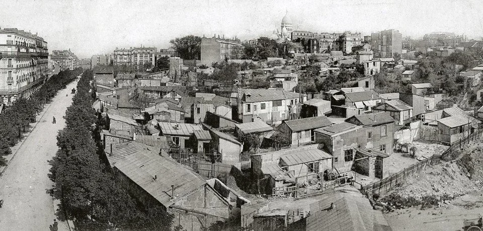 Black and white photo of a hillside slum with makeshift houses and a steep street lined with trees, and government buildings in the background.