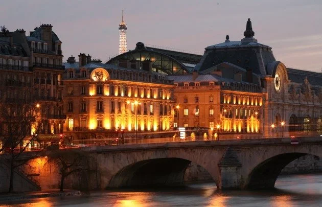 Paris cityscape at dusk with illuminated historic buildings, a bridge over the river, and the Eiffel Tower in the background.