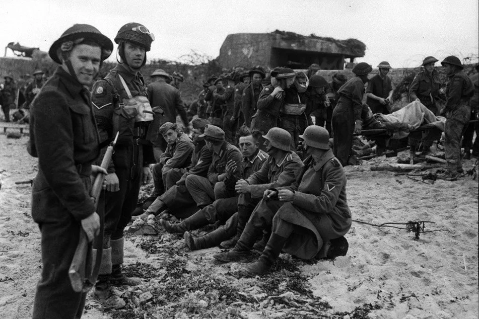 World War II soldiers resting and walking on a beach, with some sitting on the sand and others standing, near a destroyed bunker or structure.