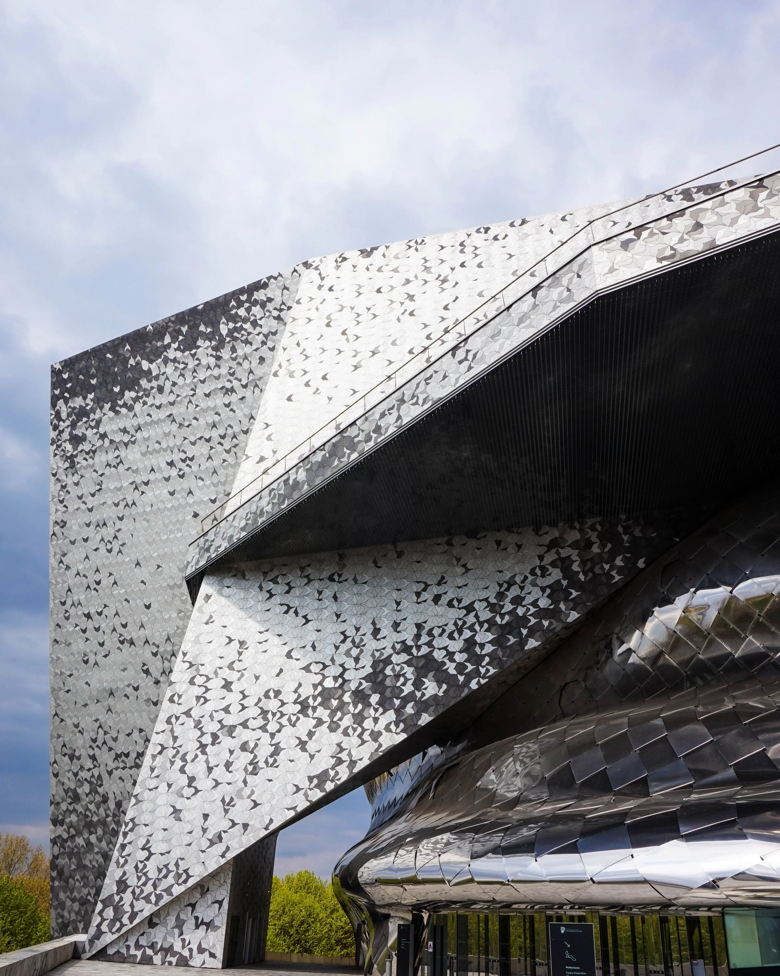 Modern building with metallic, reflective, patterned exterior and angular architecture, with trees and cloudy sky in the background.