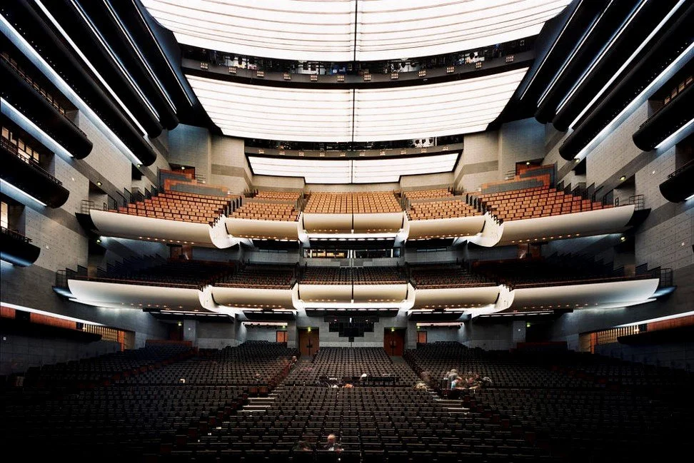 Interior of a modern theater or concert hall with multiple balcony levels, rows of empty seats, and a large illuminated ceiling.
