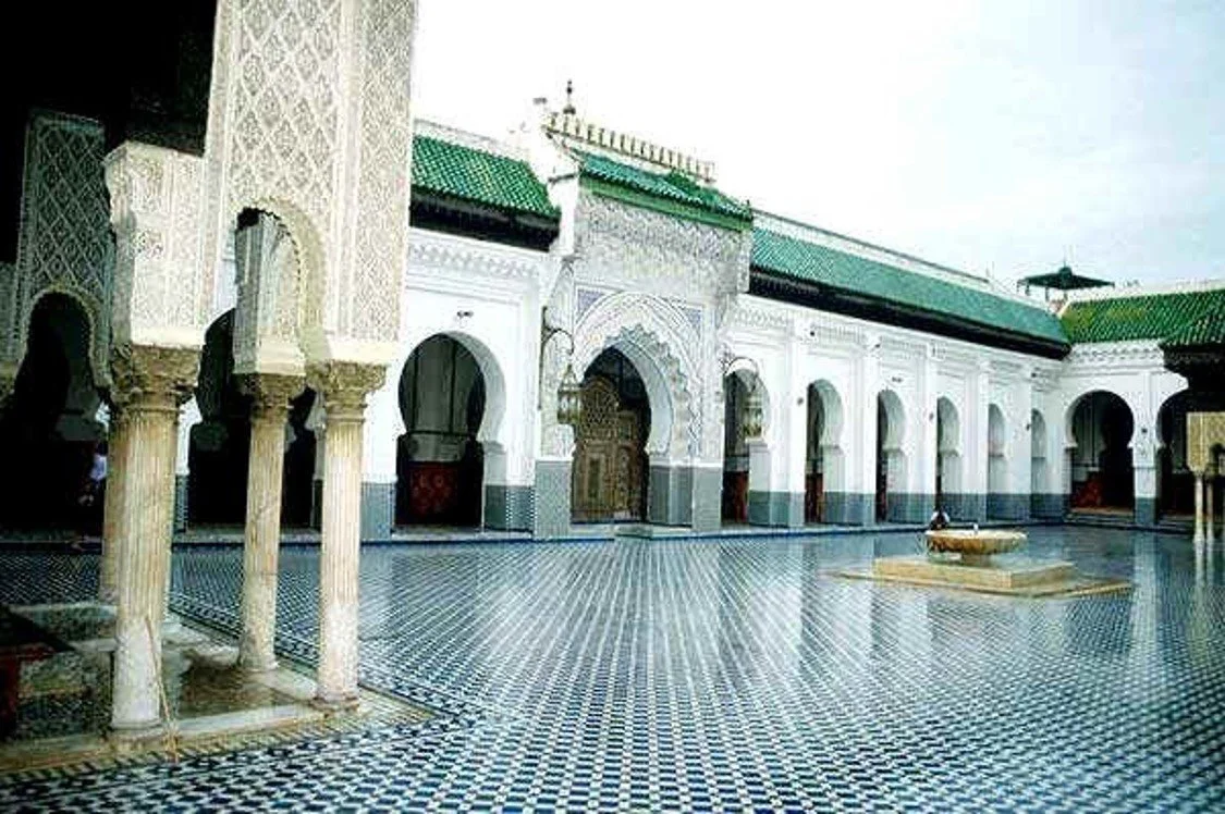 Interior courtyard of a Moroccan-style building with ornate arches, columns, green tiled roof, and a fountain in the center.