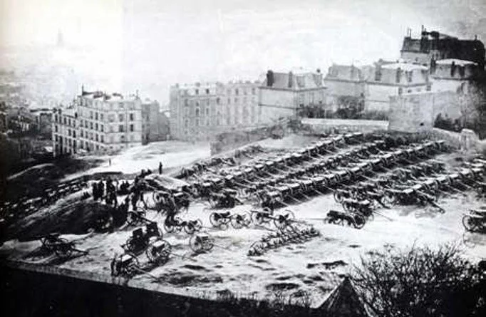 Black and white photograph of a city square with horse-drawn carriages parked in rows, some overturned, and a group of people nearby. Buildings surround the square.