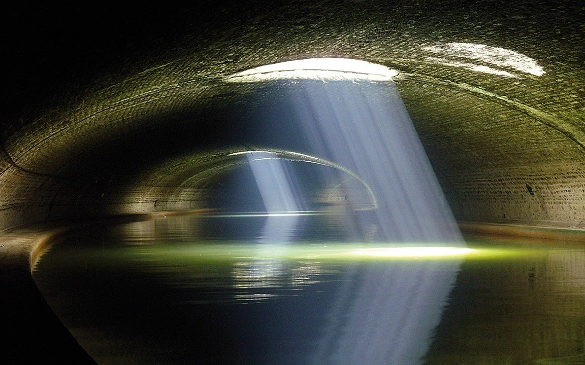 A tunnel with water at the bottom and a large opening from which sunlight streams in, creating beams of light.