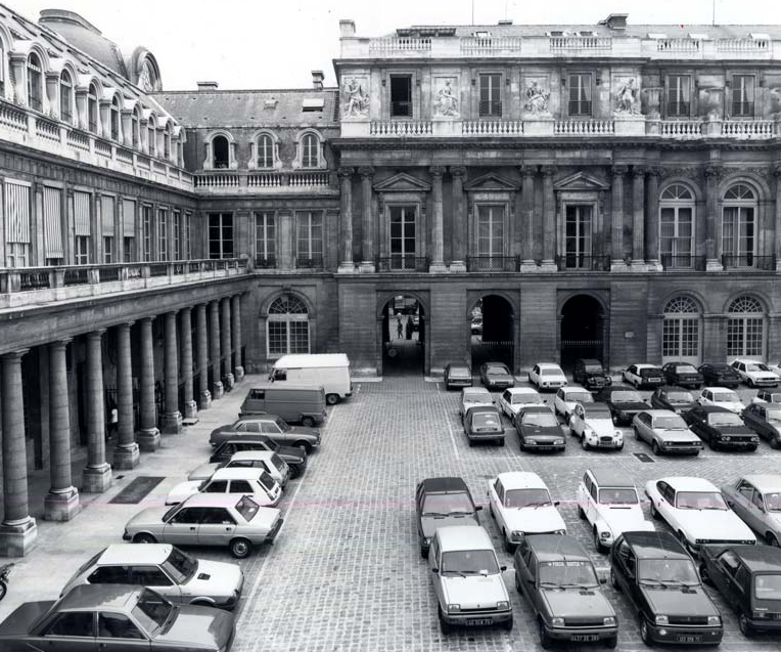 Black and white photo of an ornate historic building with a stone facade, columns, and arched windows. A courtyard with parked cars and vans is in front of the building.