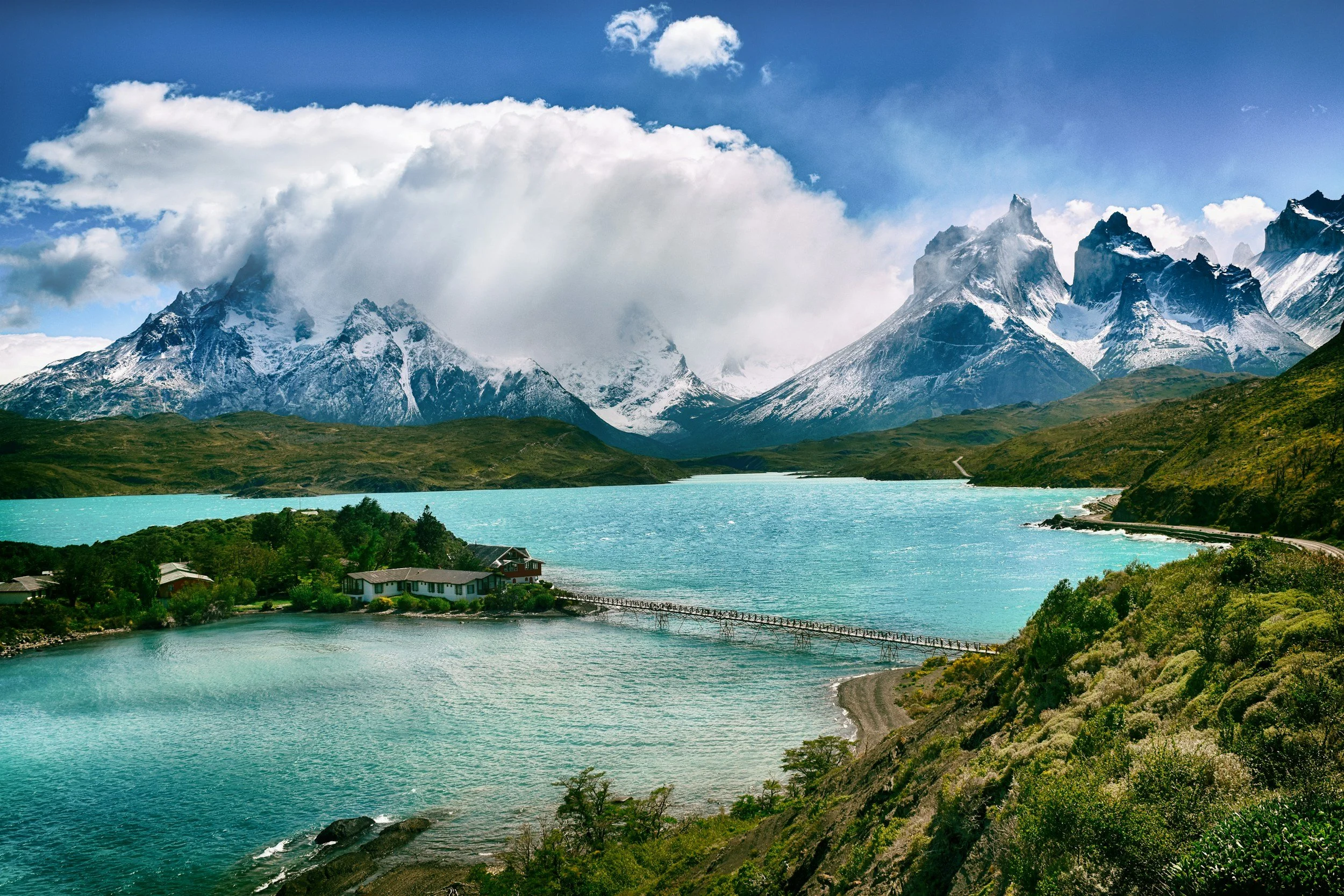Scenic view of turquoise lake surrounded by green hills and snow-capped mountains with clouds in the sky.