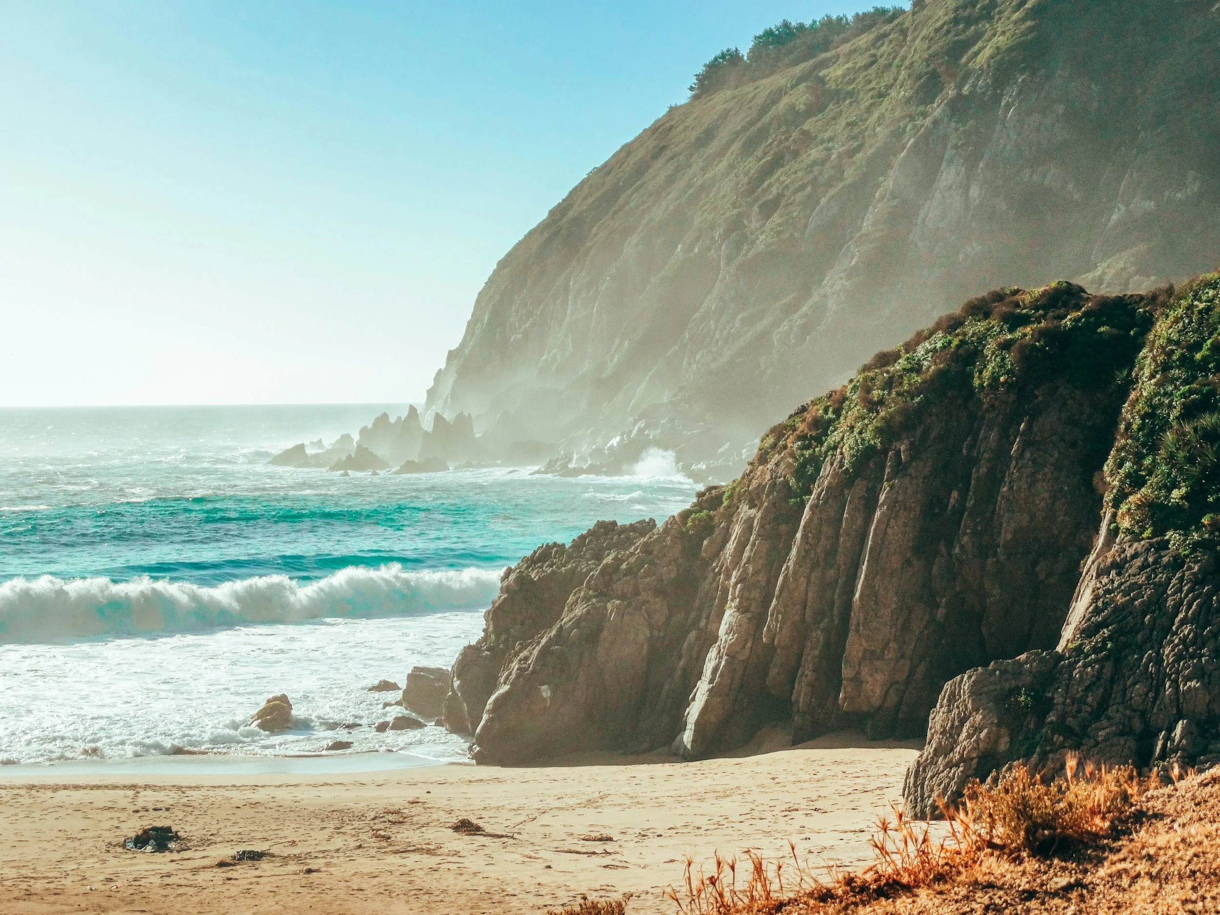 A scenic view of a rocky beach with waves crashing against the shore, large cliffs covered in greenery in the background, under a clear sky.