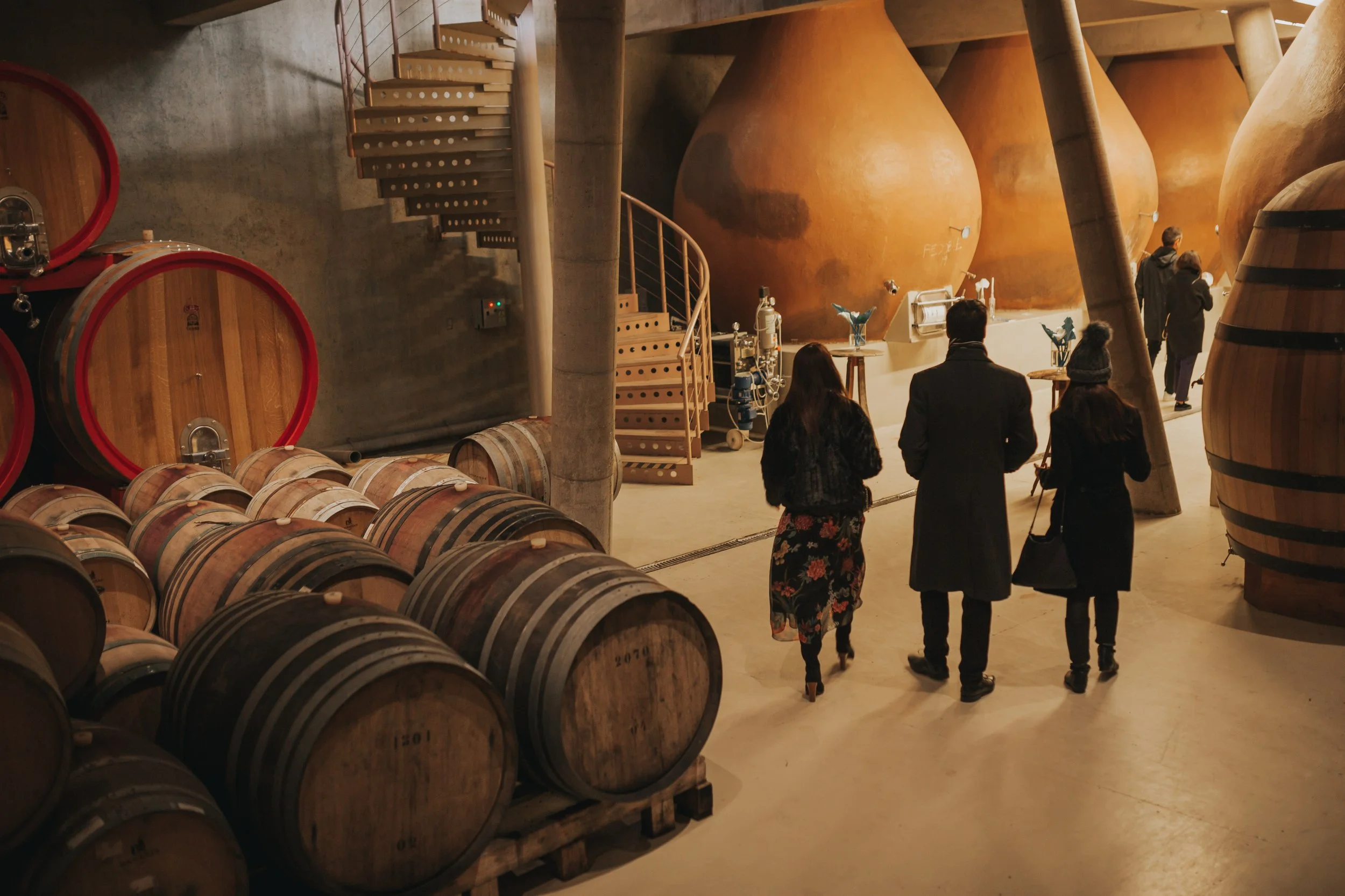 People touring a wine cellar with stacked barrels and large fermentation tanks.