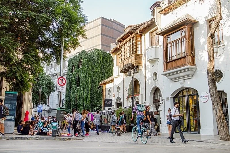 Crowd of people walking and sitting on the sidewalk in front of a white building with wooden balconies and ivy-covered trees.
