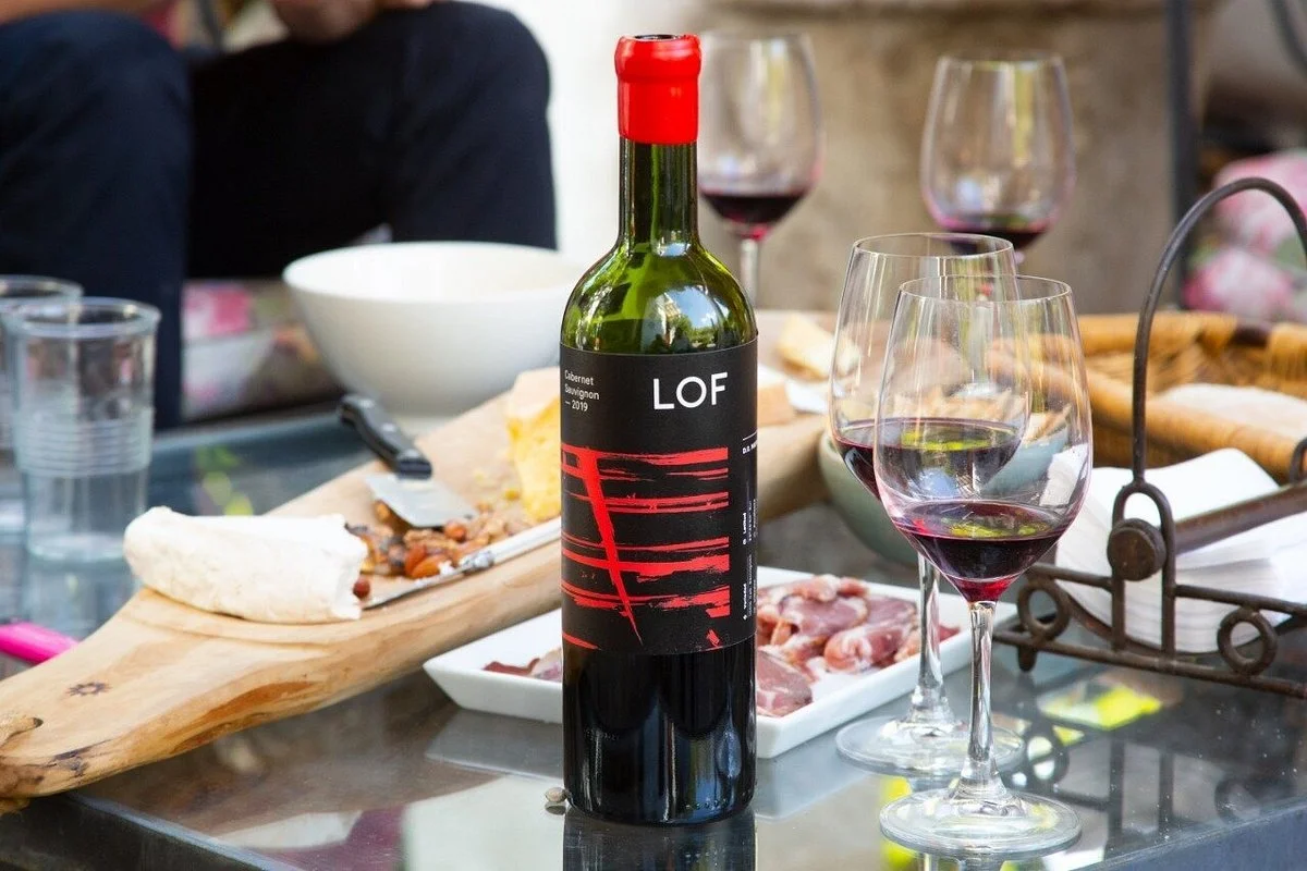 A bottle of red wine labeled 'LOF' surrounded by wine glasses filled with red wine, with a cheese board, a white bowl, and some food on a glass dining table.