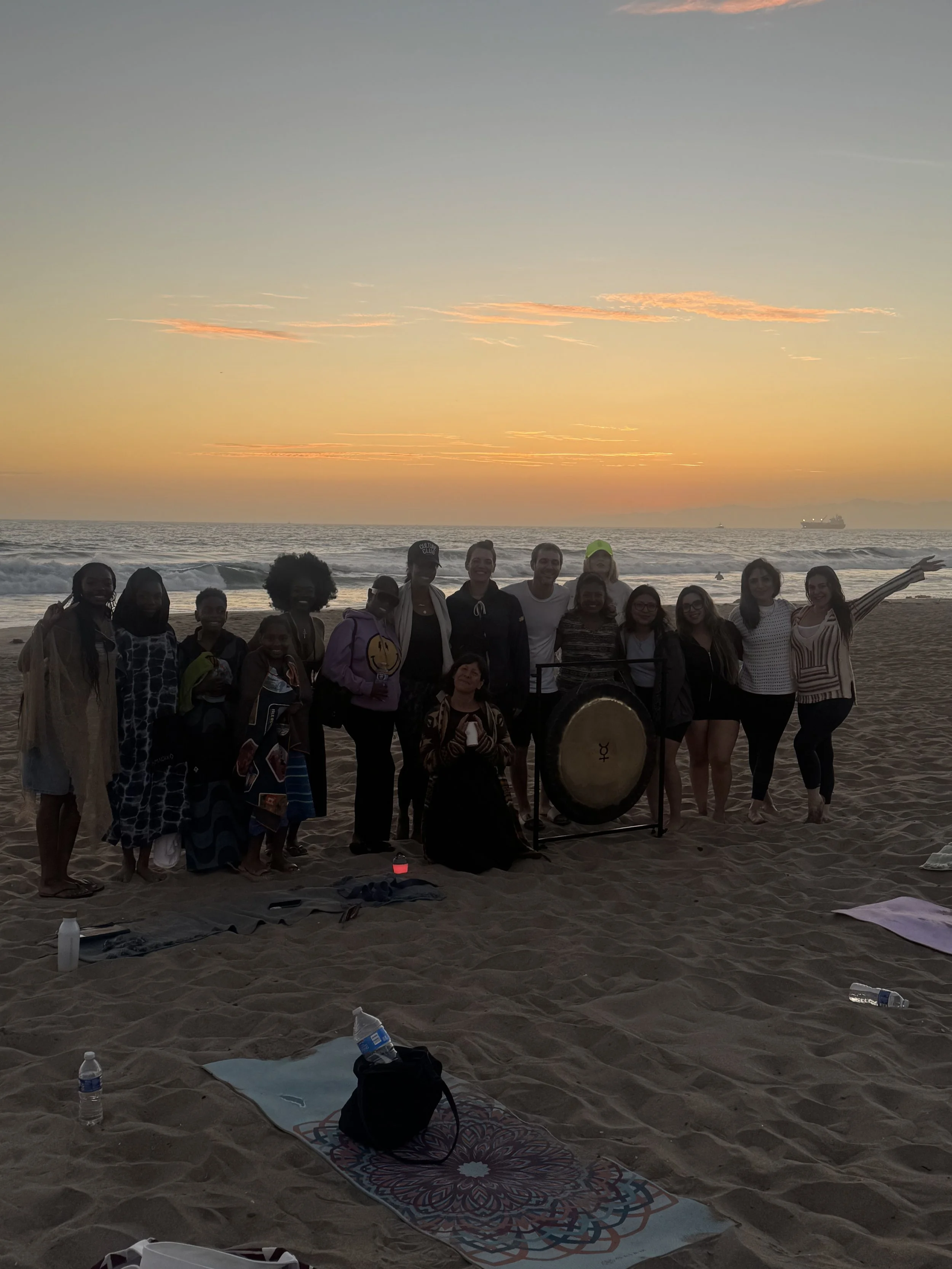Group of people standing on a beach during sunset, with ocean waves and two ships in the background, some holding drumsticks and a large drum in the center.