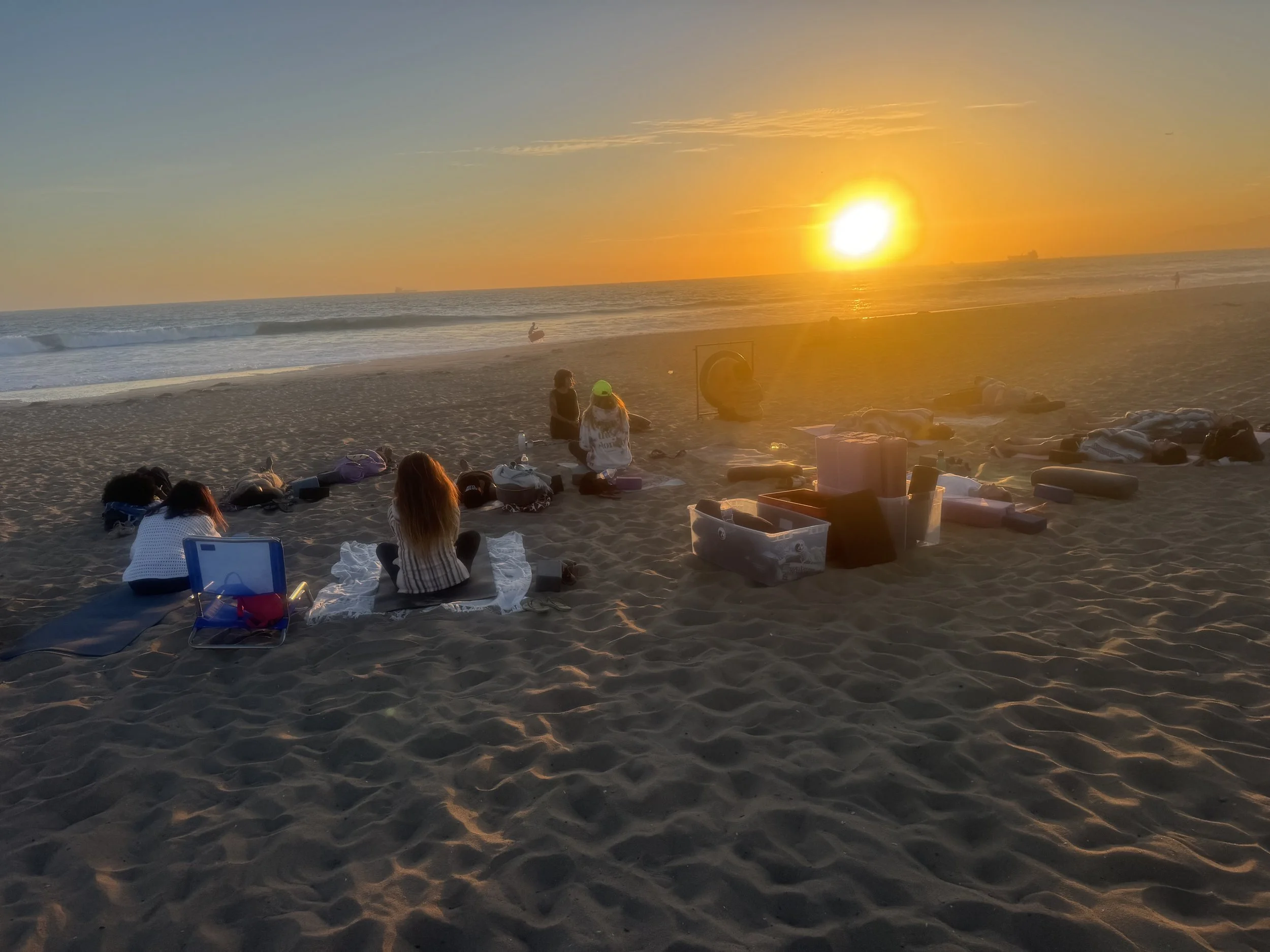 People sitting on the sand at the beach during sunset, with the ocean waves in the background and a ship visible on the horizon.