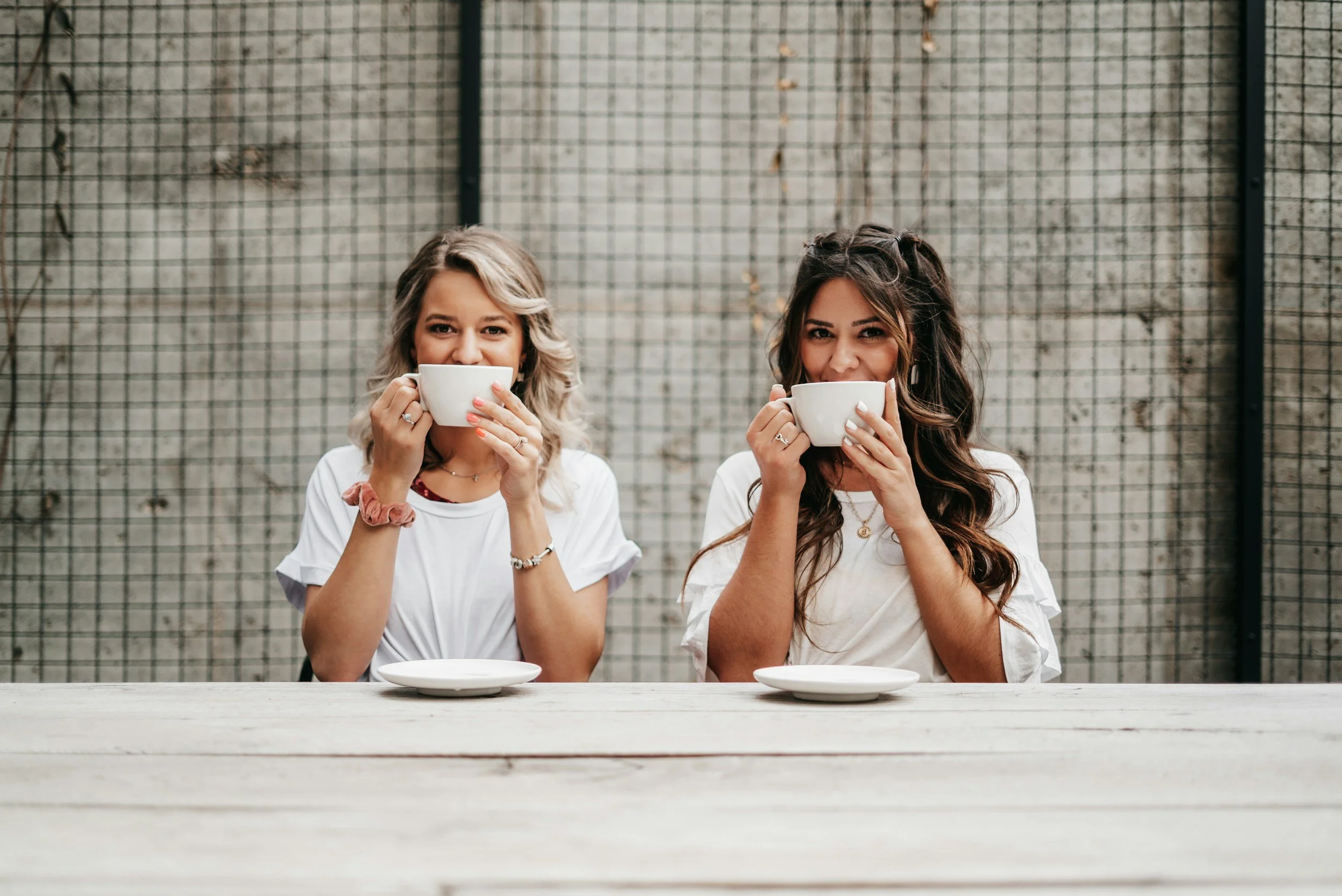 Two women sitting at a table, holding coffee cups near their faces, smiling, with a light-colored wooden table and a wire fence in the background.
