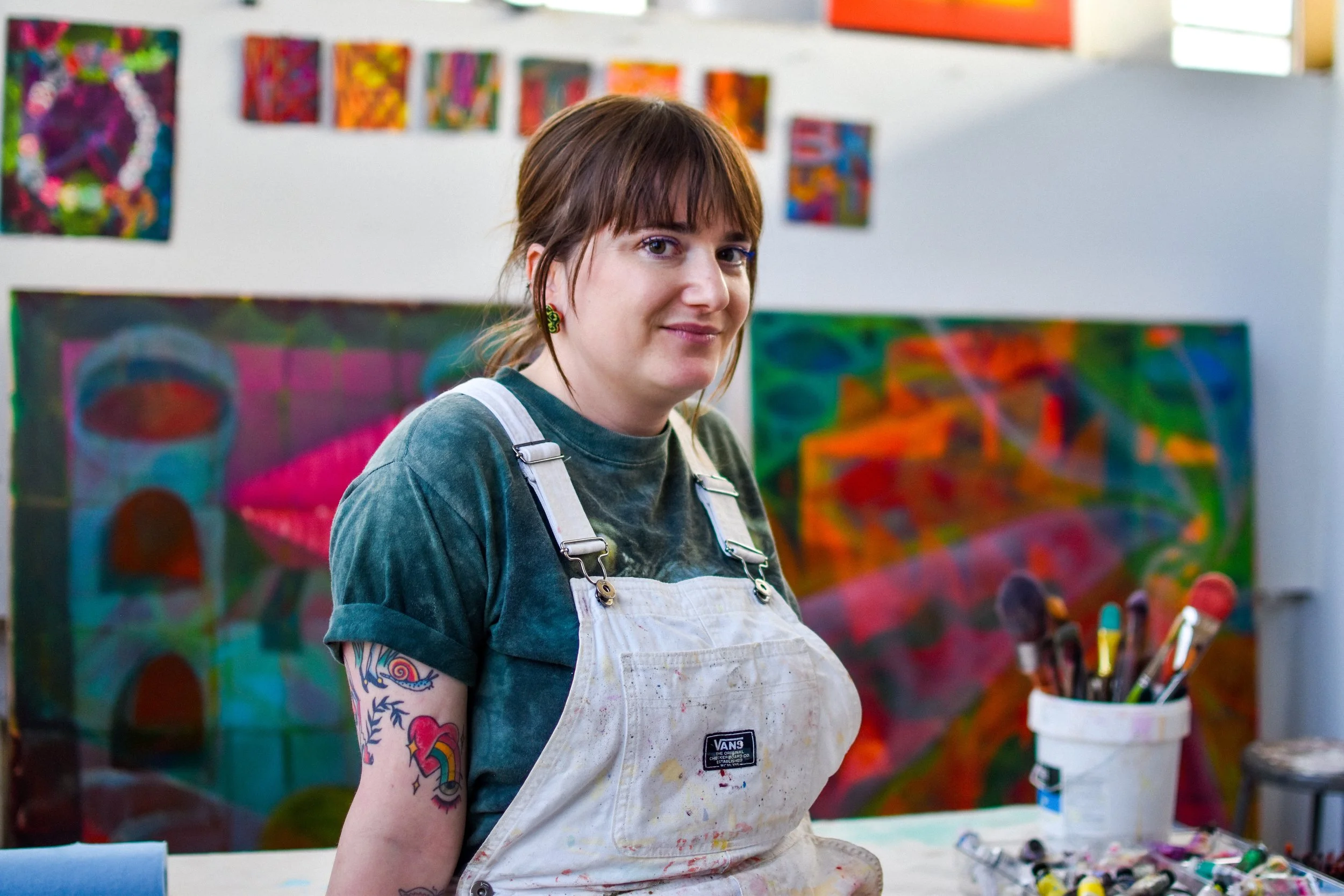 A woman with short brown hair and tattoos on her left arm, wearing a dark green shirt and white apron, standing in an art studio with colorful abstract paintings behind her and paintbrushes in a container on a table.