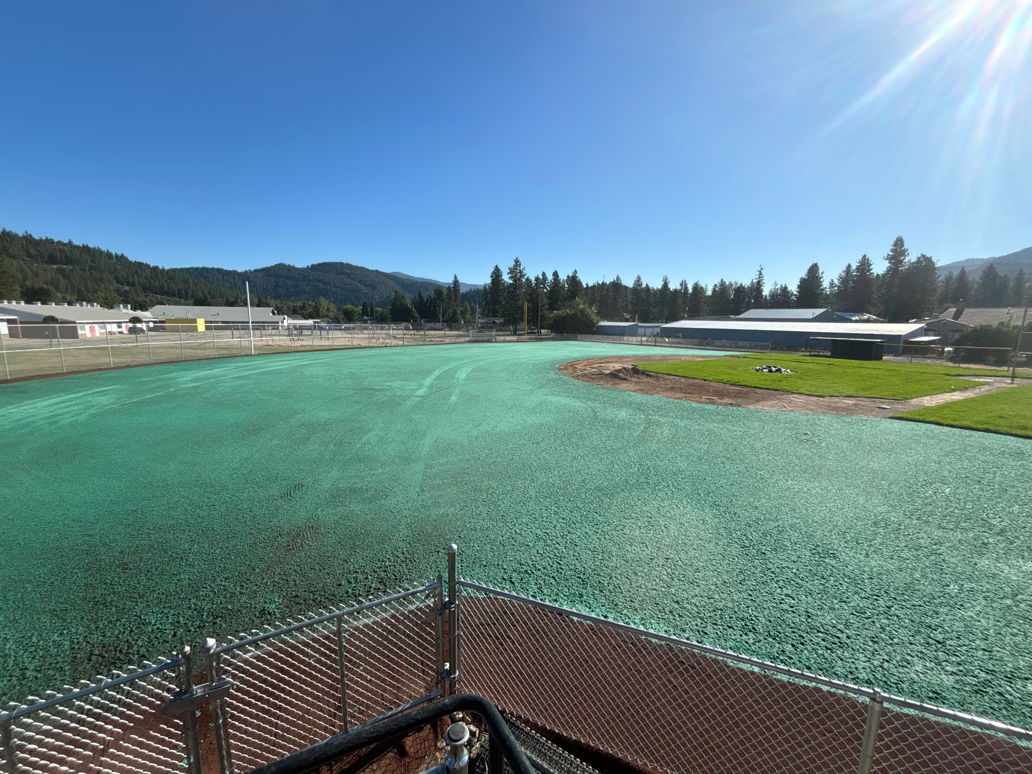 Fenced area with green hydroseeding on a sports field, surrounded by buildings and trees, under a clear blue sky.